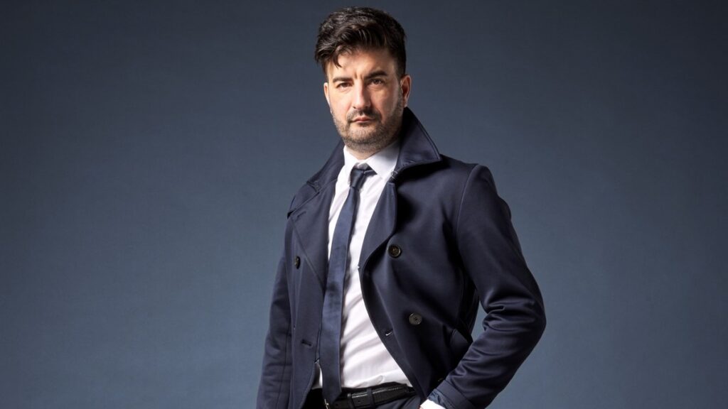Portrait professionnel de Sam Brocal, consultant en marketing digital chez Symphon’e-Communication, debout en costume bleu marine sur fond bleu-gris. L’image accompagne un article sur un cas client Odoo Online, le tracking e-commerce, Meta Ads et le server-side tracking.